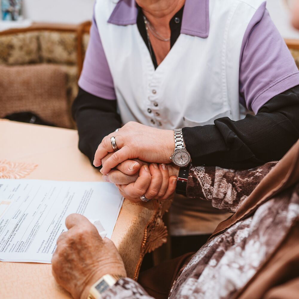 Eine Pflegerin hält die Hand einer Seniorin und hilft ihr beim Anträge ausfüllen.